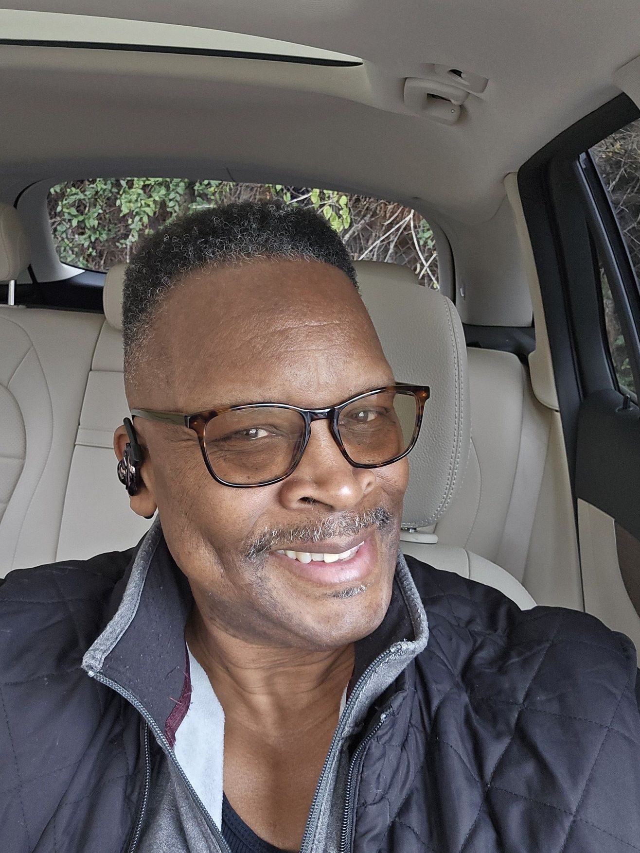 Man in car, wearing glasses, smiling. Black jacket, light-colored interior, outdoor background.