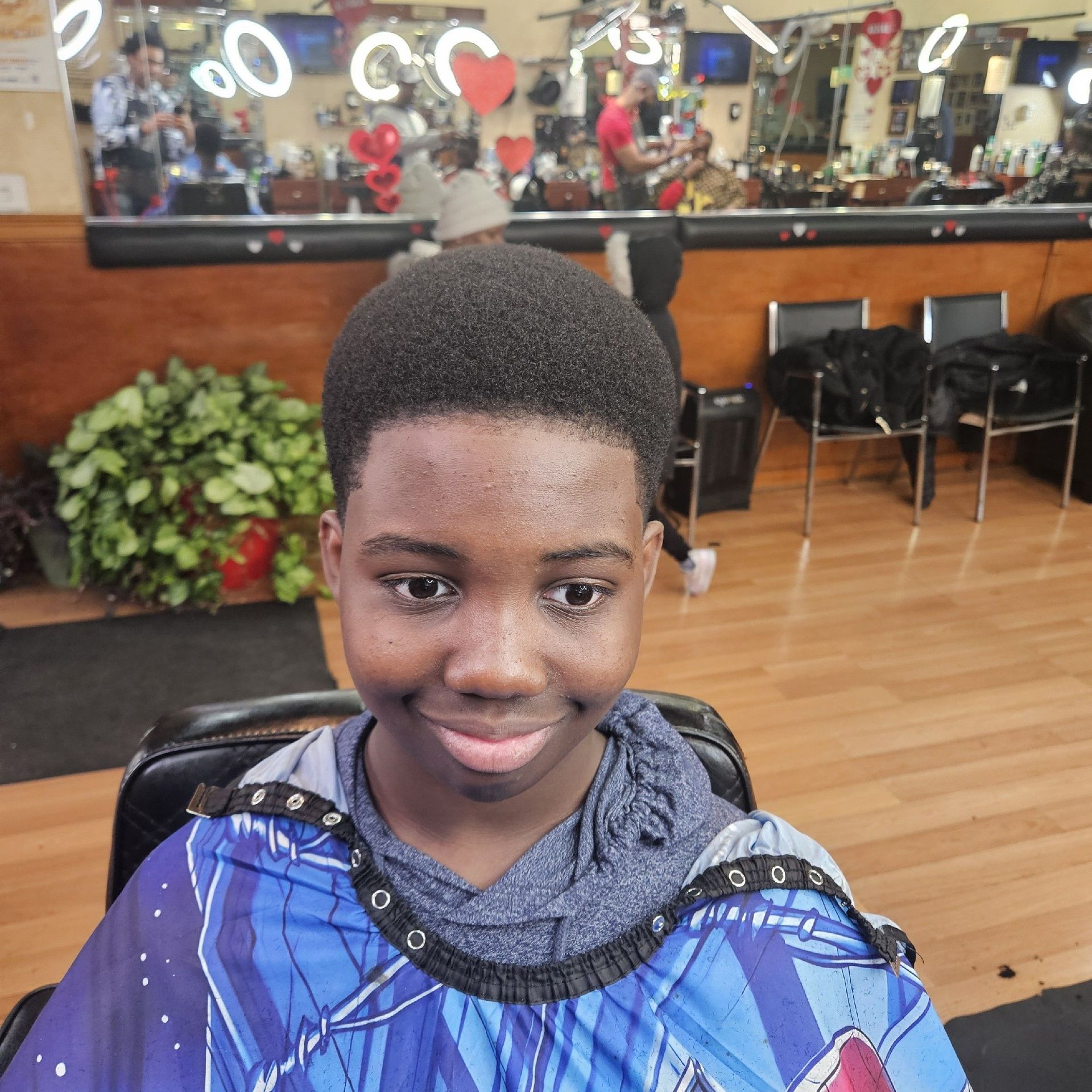 Boy in a barber shop, smiling after haircut. He is wearing a cape. Shop has a mirror and other people.