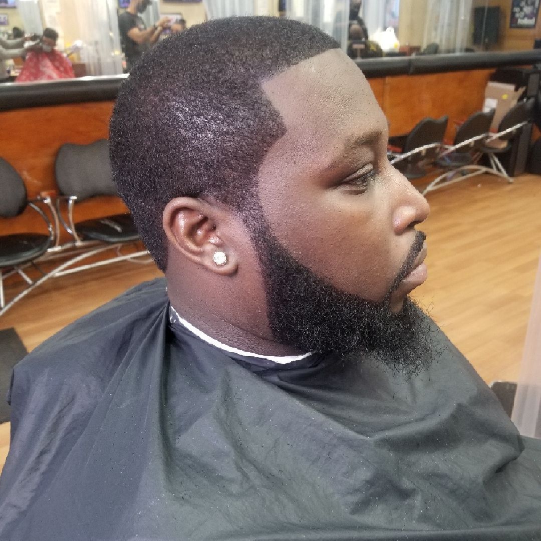 Man with a dark, short haircut and full beard, wearing a black barber's cape at a barbershop.