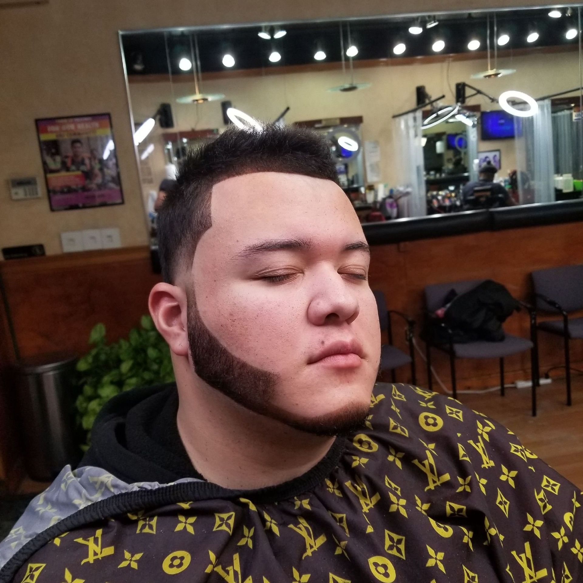 Man with a new haircut and beard sits in a barber shop, wearing a protective cape.
