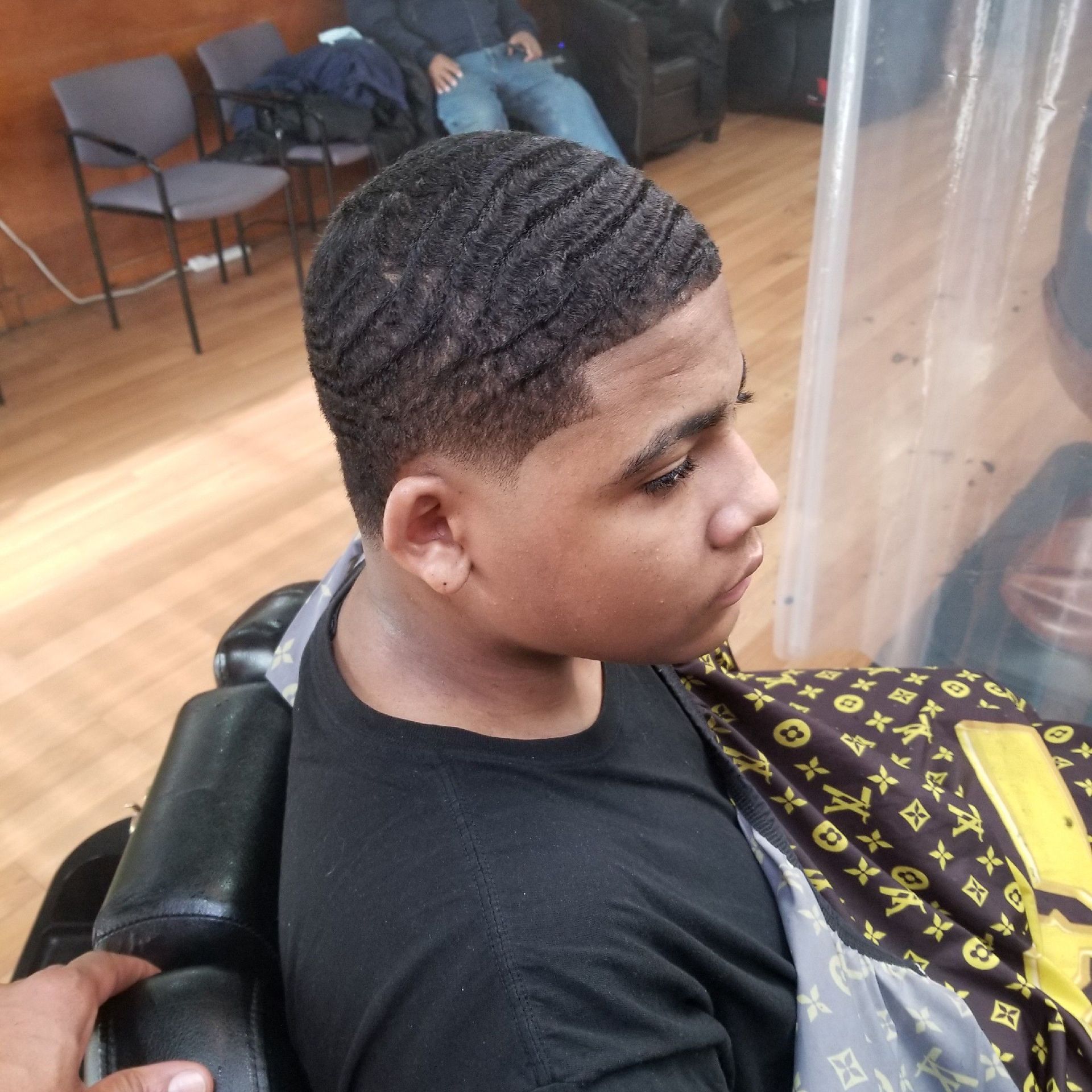 A young person with a wave haircut sitting in a barber chair. They are wearing a black shirt and looking to the side.