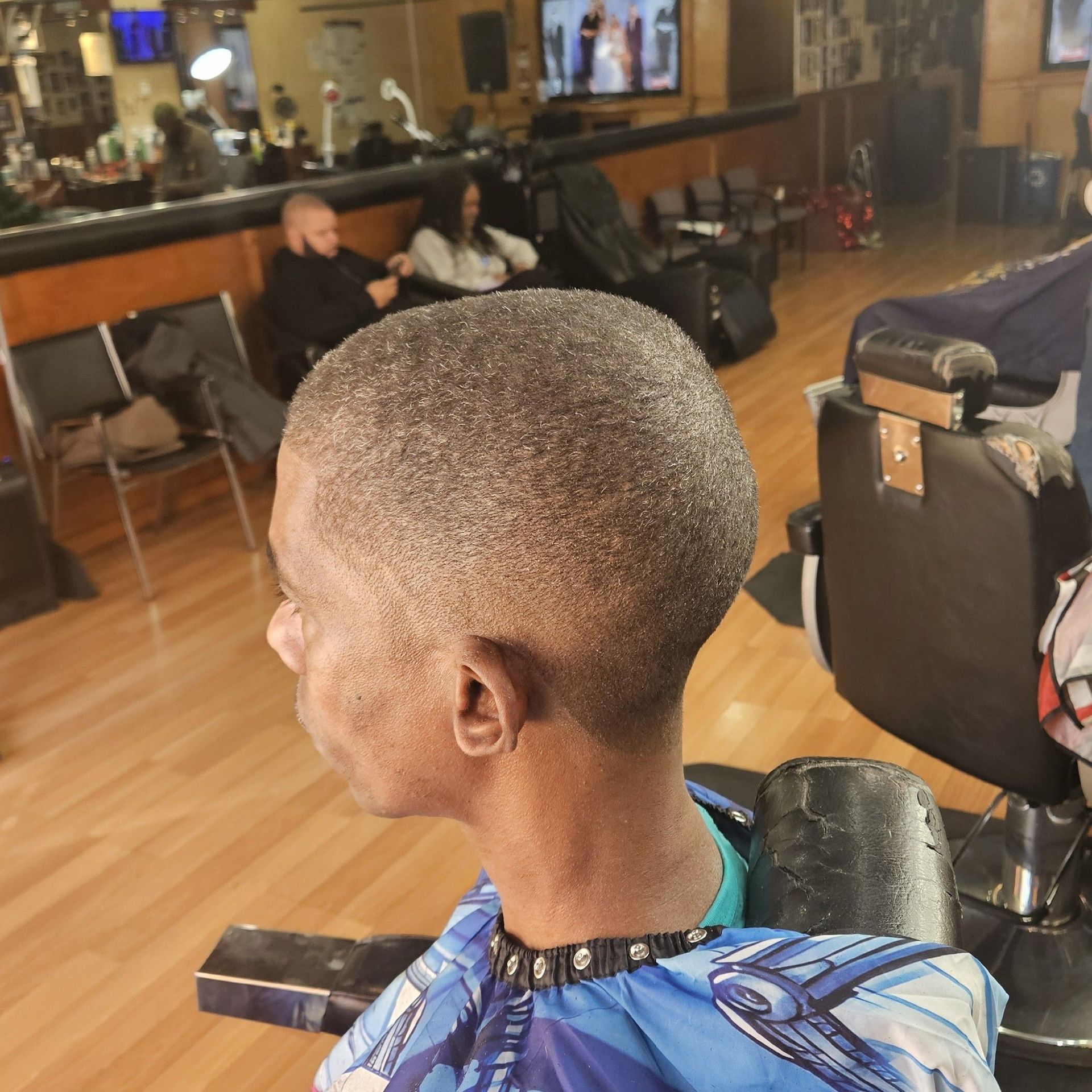 Man with short haircut at a barbershop, wearing a blue cape. Wooden floor and other patrons are in the background.