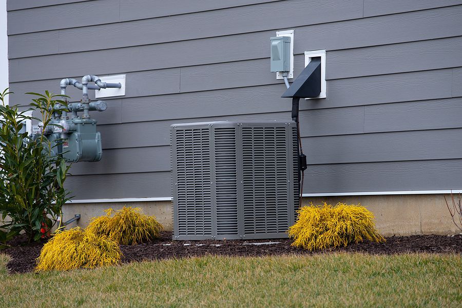 Outdoor home utility equipment beside a gray house wall, with vents and yellow shrubs in front.