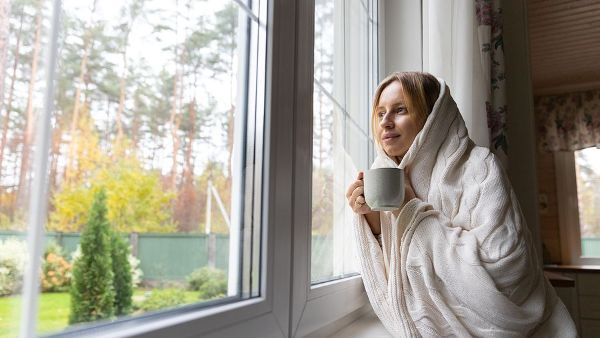 A woman wrapped in a blanket is sitting on a window sill holding a cup of coffee.