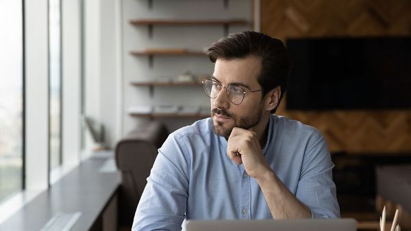 A man is sitting at a table with a laptop and looking out the window.