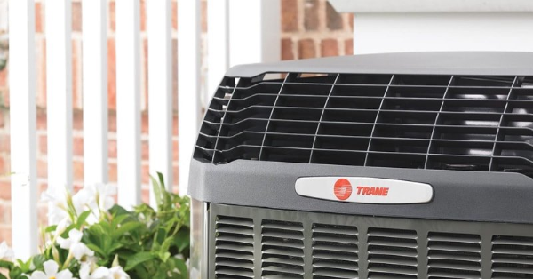 Gray air conditioning unit with the Trane logo, outdoors near white picket fence and greenery.