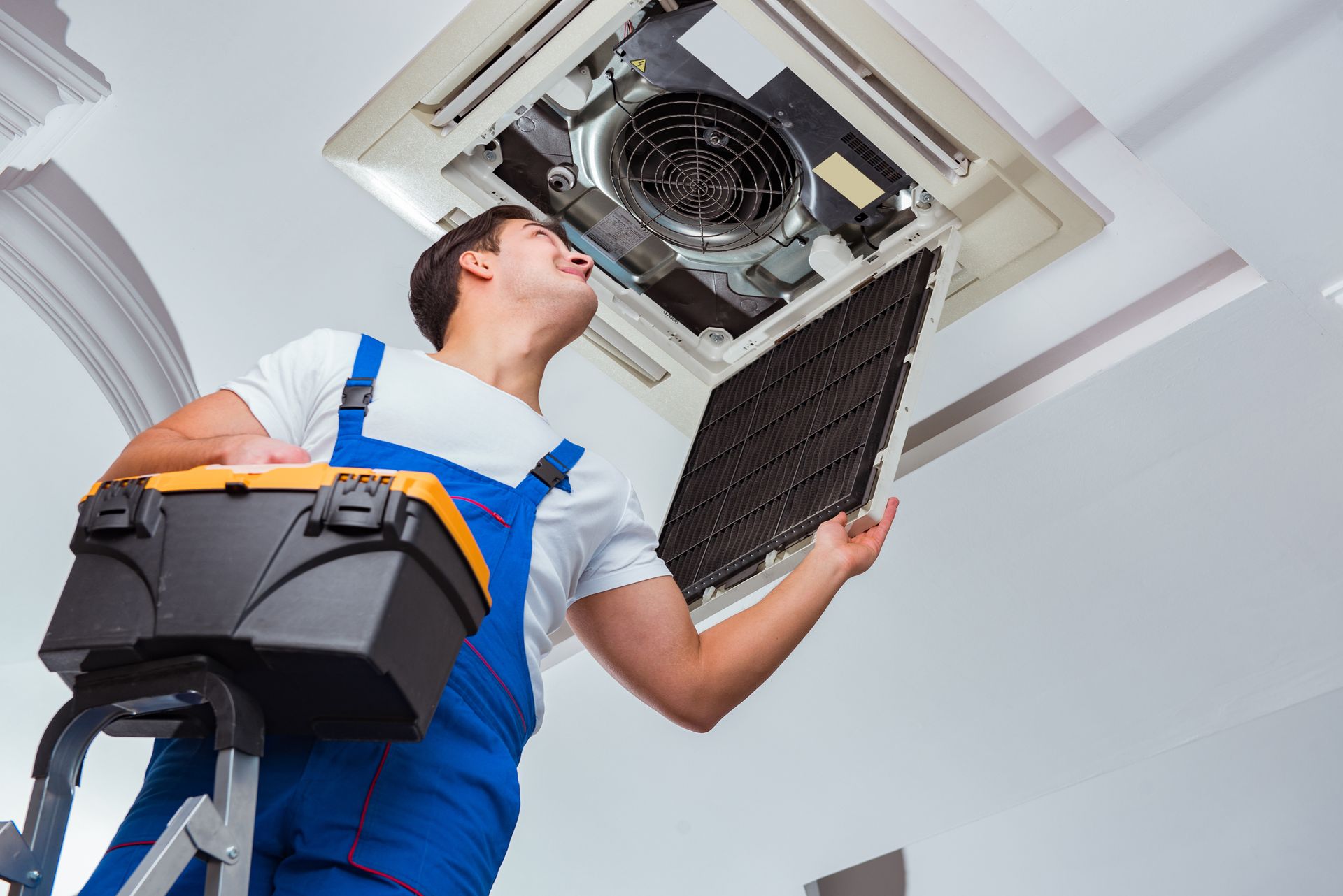 HVAC technician on a ladder, inspecting ceiling air conditioner, holding a filter and toolbox.