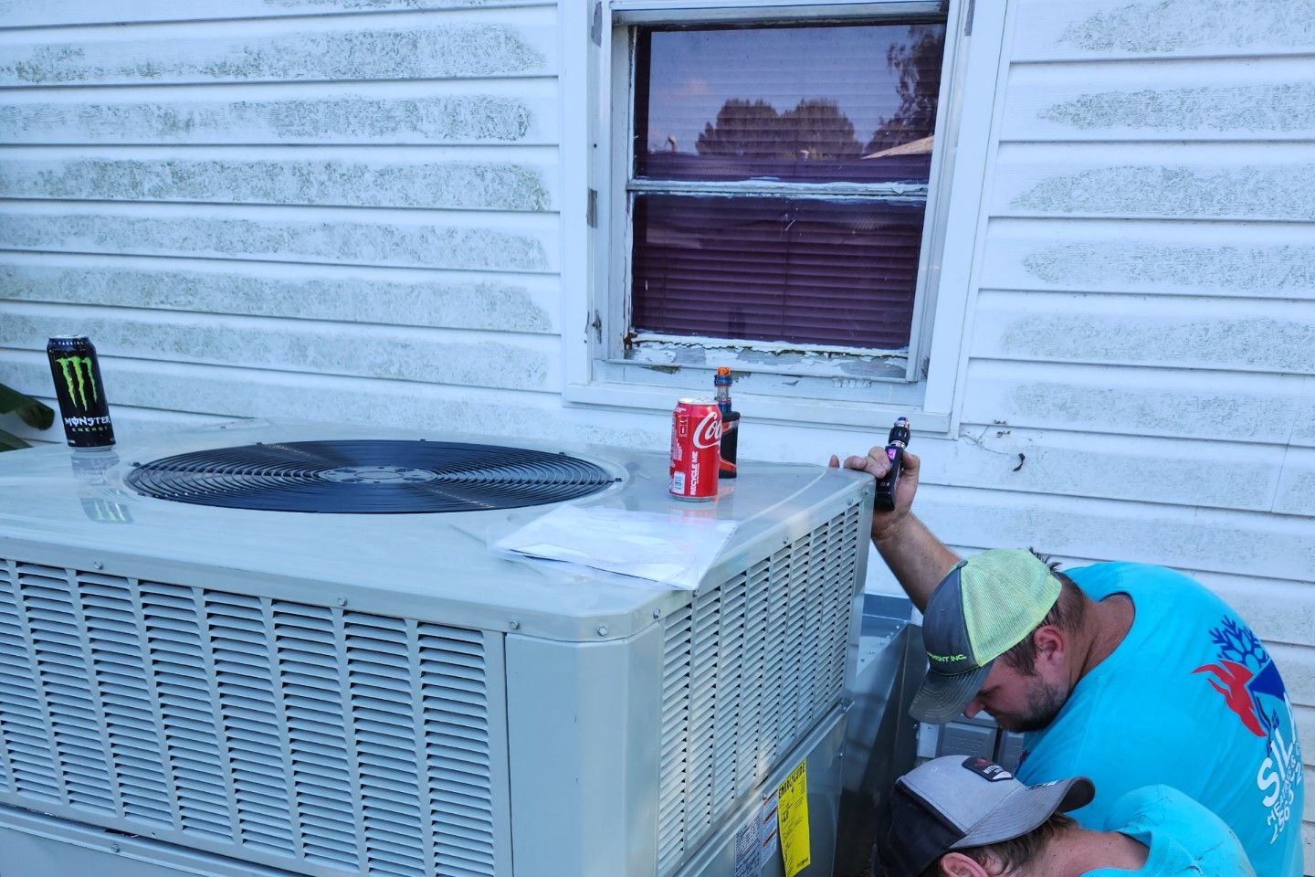 Two men in work shirts repair an air conditioning unit next to a house; drinks sit on top.