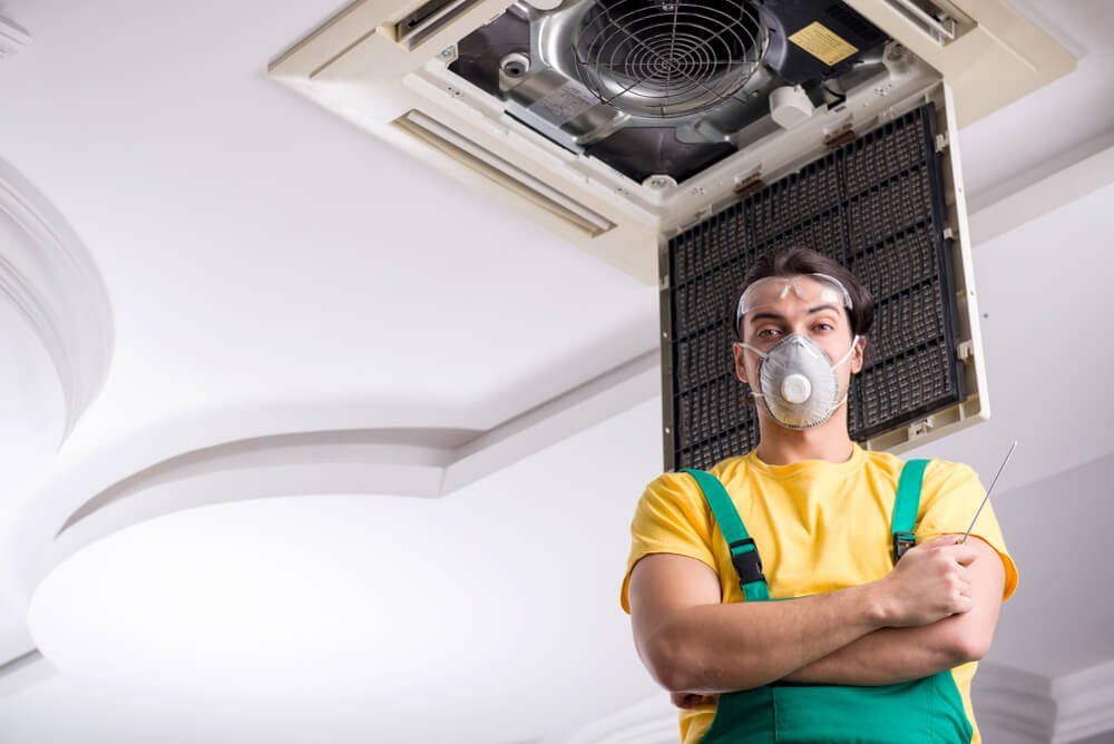 HVAC technician in yellow shirt and green overalls, wearing a respirator, working on a ceiling air vent.