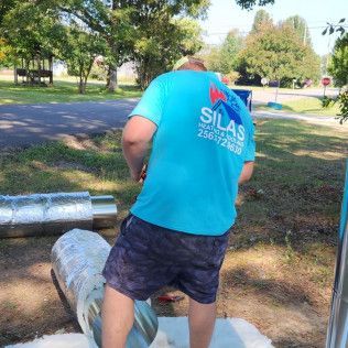 Man in a blue shirt with logo cutting metal ductwork outdoors.