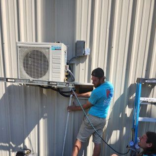 HVAC technicians installing an AC unit on a corrugated metal building exterior.