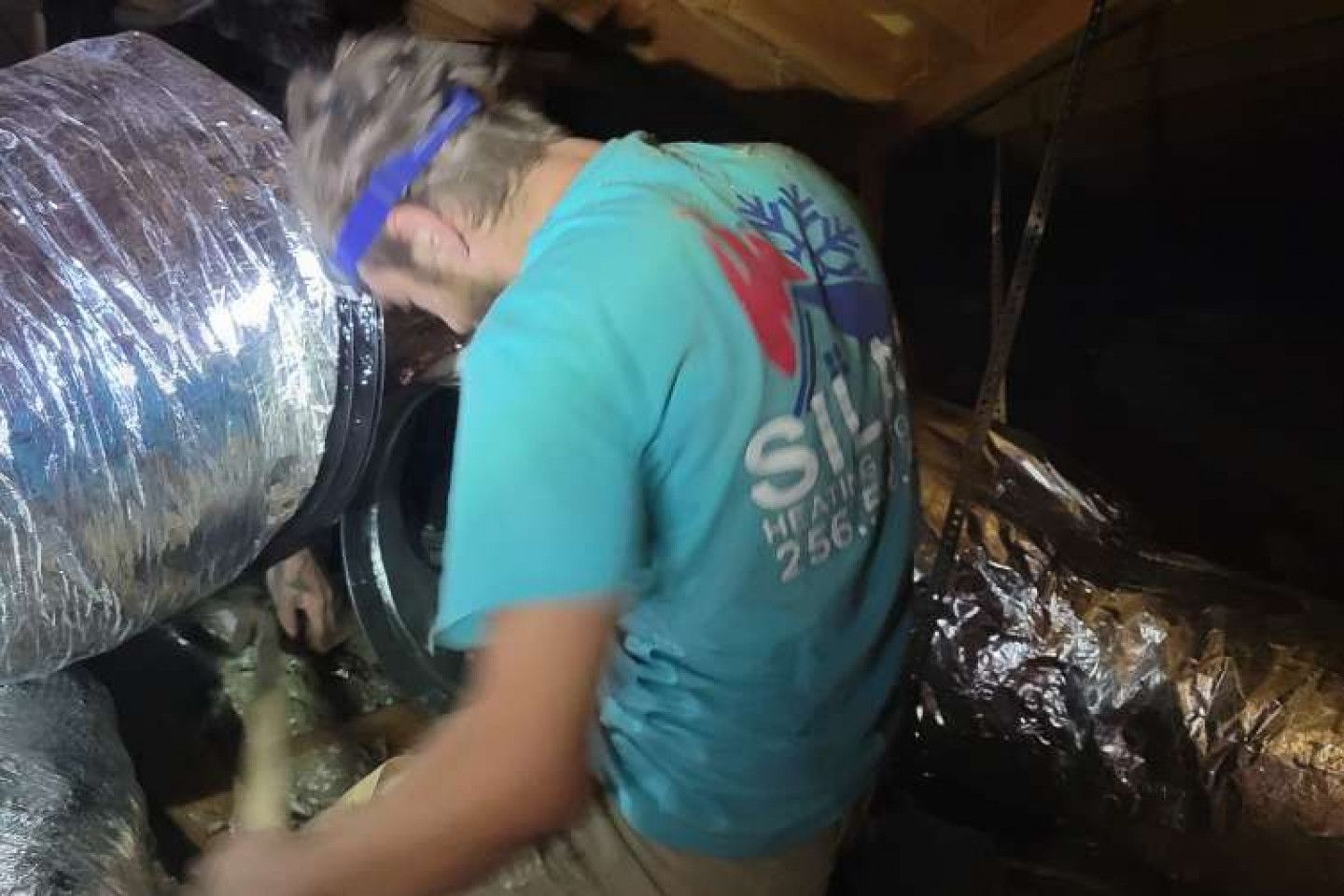 HVAC worker in a dark crawlspace inspects ductwork, wearing a blue headband and shirt with company logo.