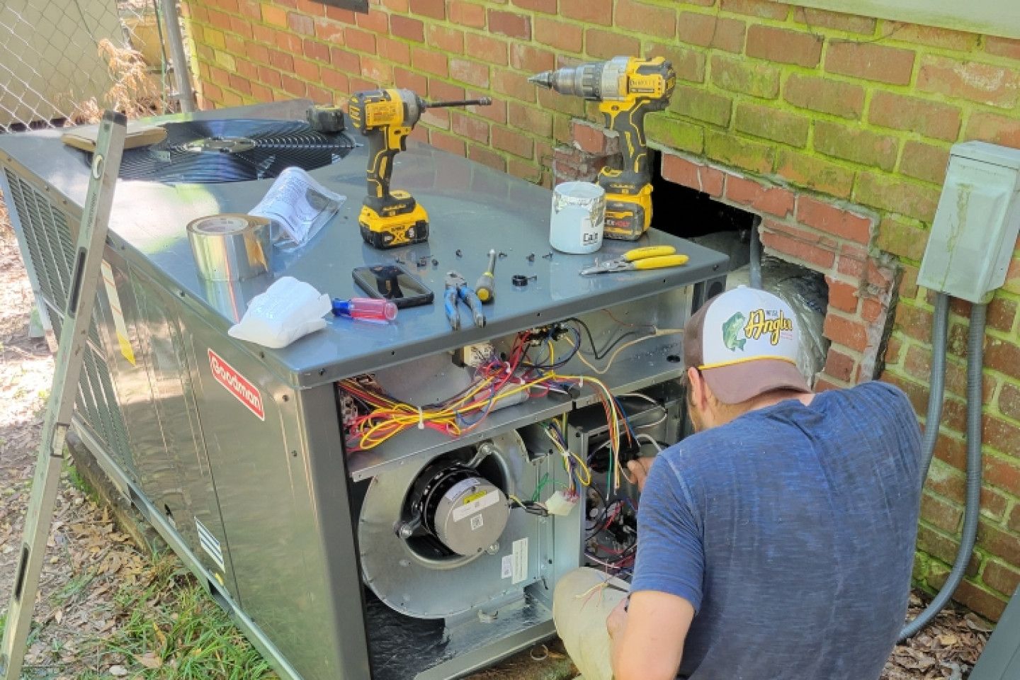 HVAC technician repairing furnace outdoors near a brick wall, tools scattered on top, sunny day.