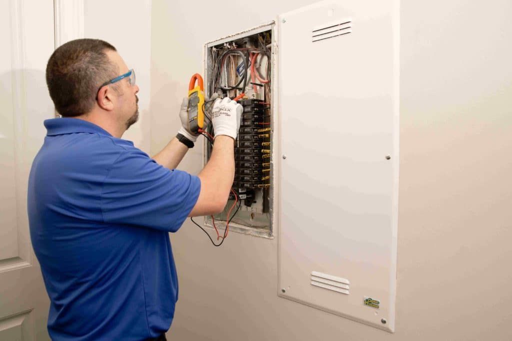 Electrician using a clamp meter on a circuit breaker panel. Indoors, white wall.