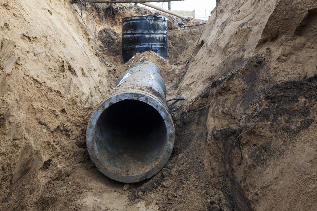 A large pipe in a trench, connected to a black cylindrical structure. Sandy soil surrounds.