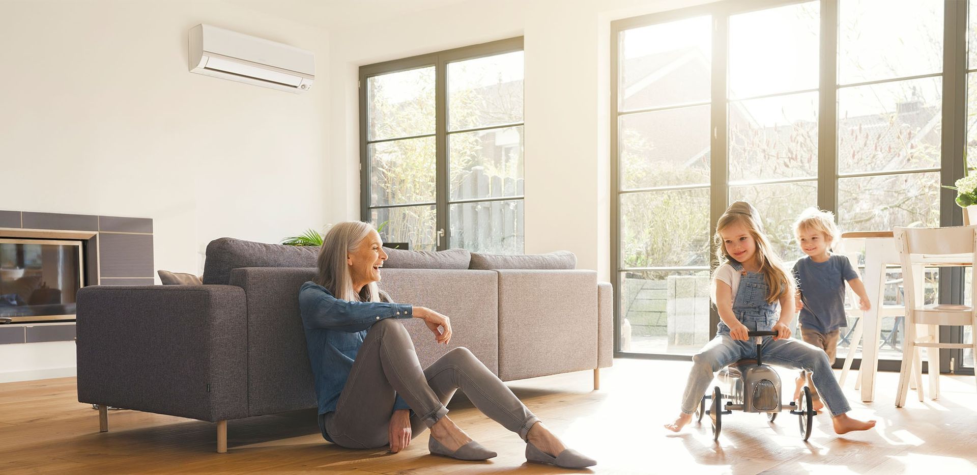 An older adult sits near a couch as two children play indoors. Sunlight streams in through windows.