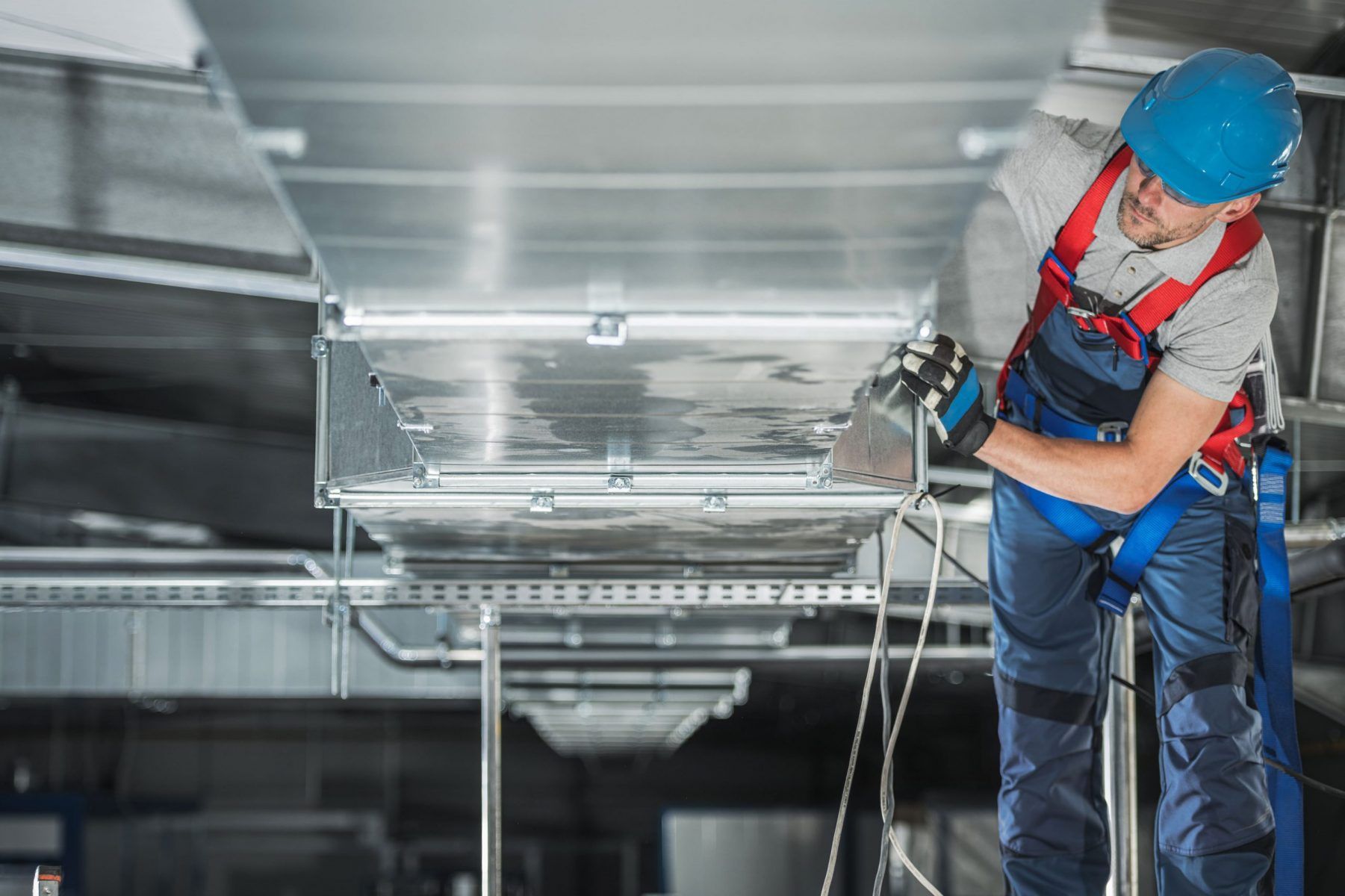 HVAC technician in blue jumpsuit and helmet working on overhead ductwork, wearing safety harness.