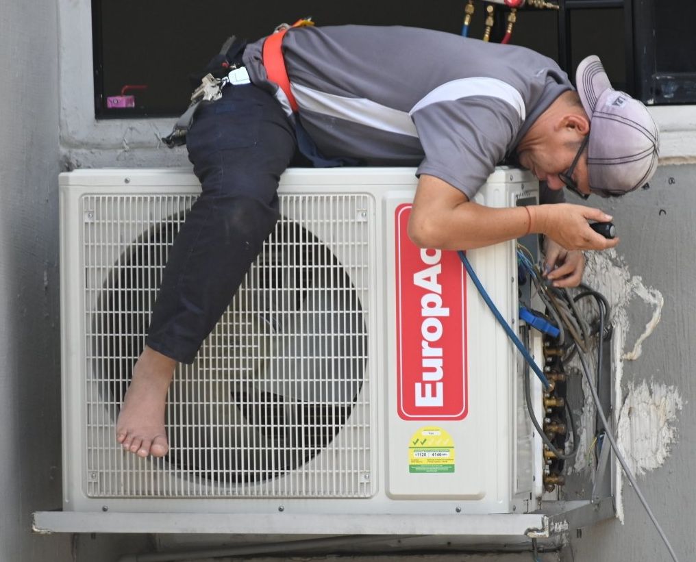 Person working on an air conditioning unit. He is lying on top with a tool in hand. Outdoors.