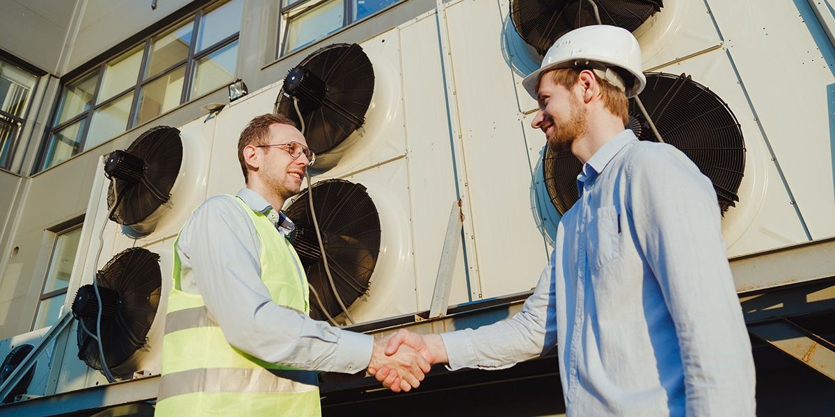 Two men shaking hands in front of industrial air conditioning units. One man wears a hard hat and the other a safety vest.