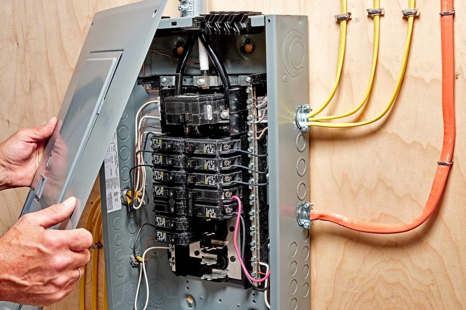 Person opening an electrical panel, revealing circuit breakers and wires. Orange and yellow cables run outside.