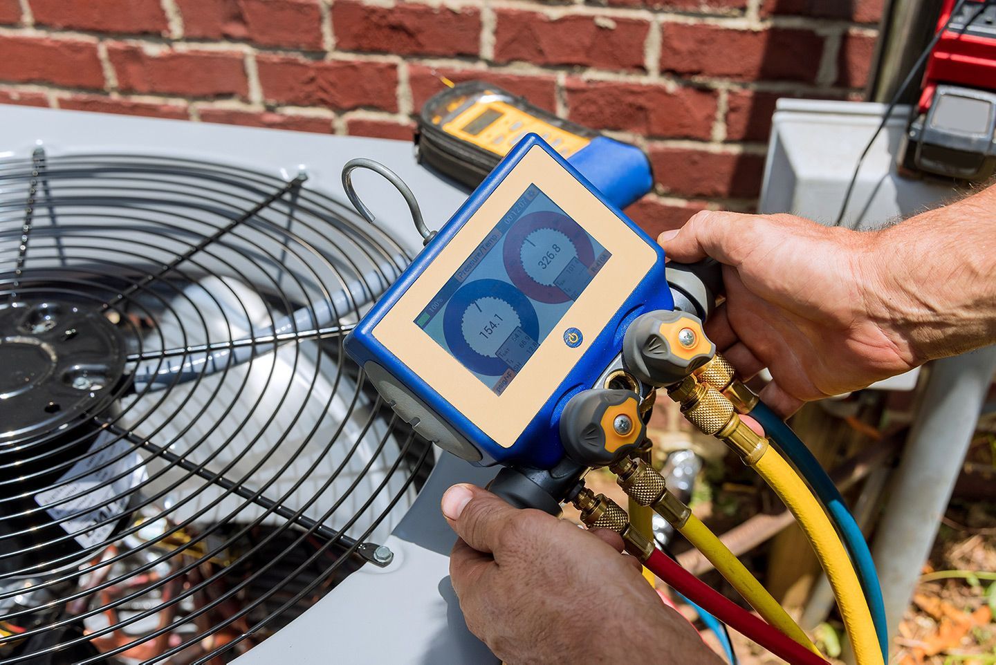Hands connecting gauges to an air conditioner unit near a brick wall. Yellow, blue, and red hoses are connected.