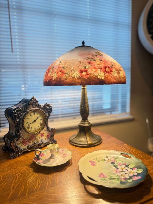 Table with floral lamp, clock, and plates in front of a window with blinds.