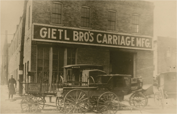 Gietl Bros' Carriage Mfg. building with horse-drawn carriages parked in front, a man stands nearby.
