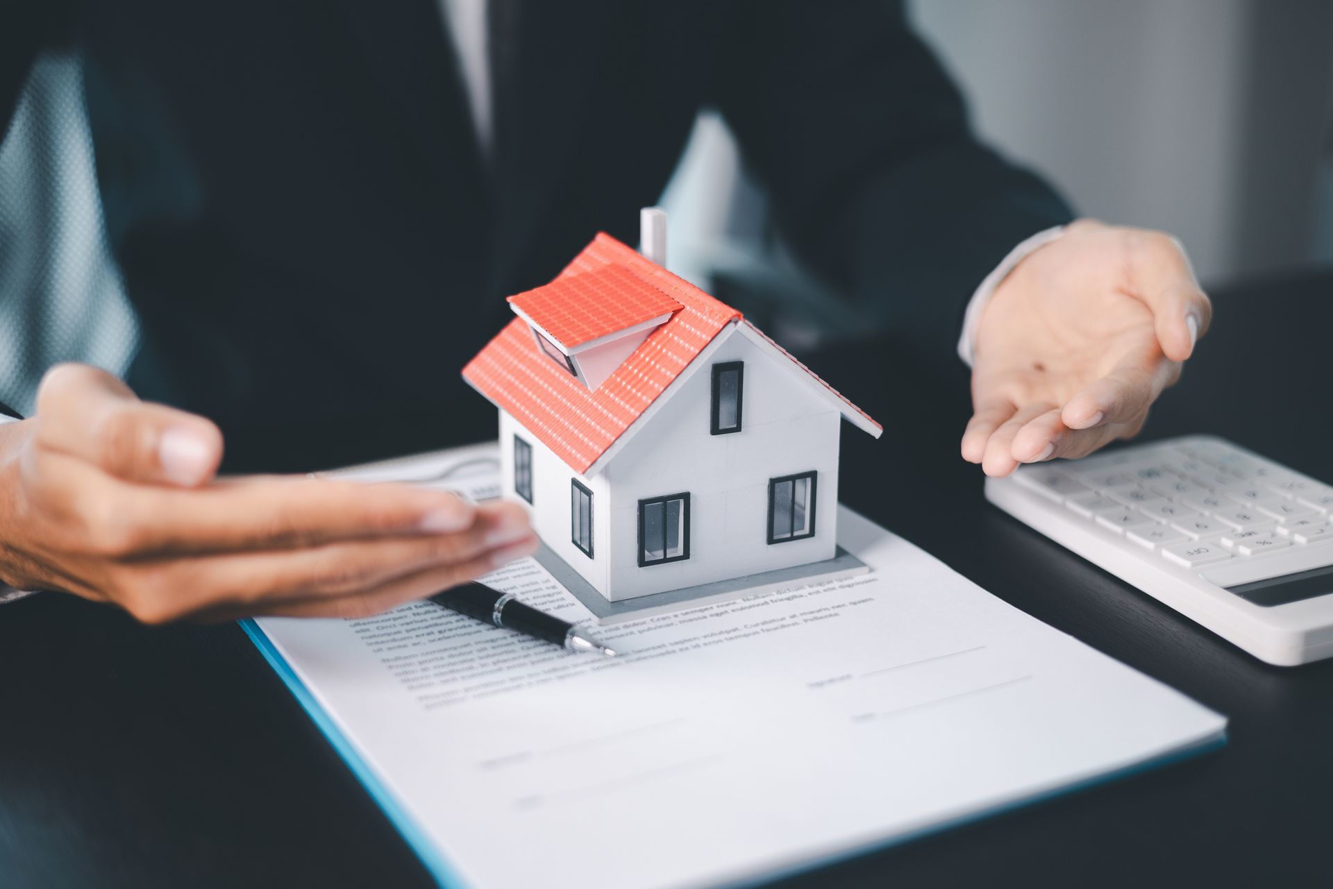 Person presenting a miniature house on top of paperwork, next to a calculator, symbolizing a real estate transaction.