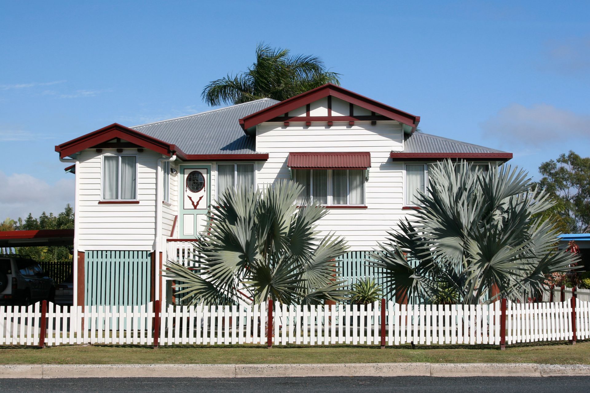 Two-story white house with a picket fence, blue sky, and silver fan palms in front.
