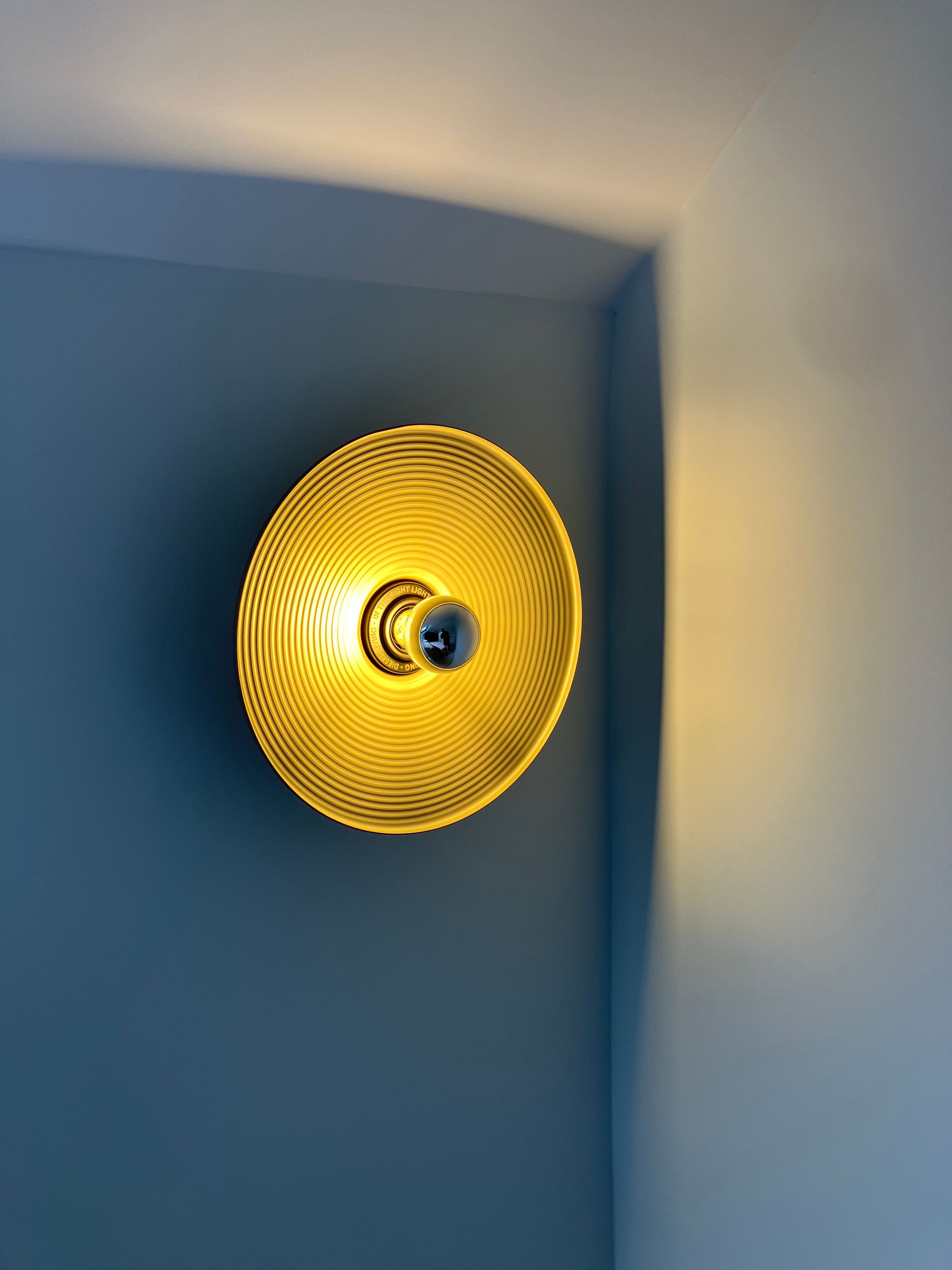 Yellow, patterned wall sconce illuminates a corner. The fixture is circular with concentric ridges.