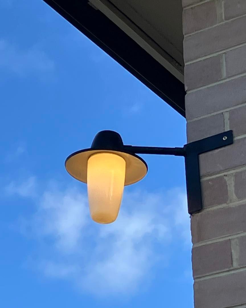 Black outdoor wall-mounted lamp with a white cylindrical shade against a blue sky, mounted on a brick wall.