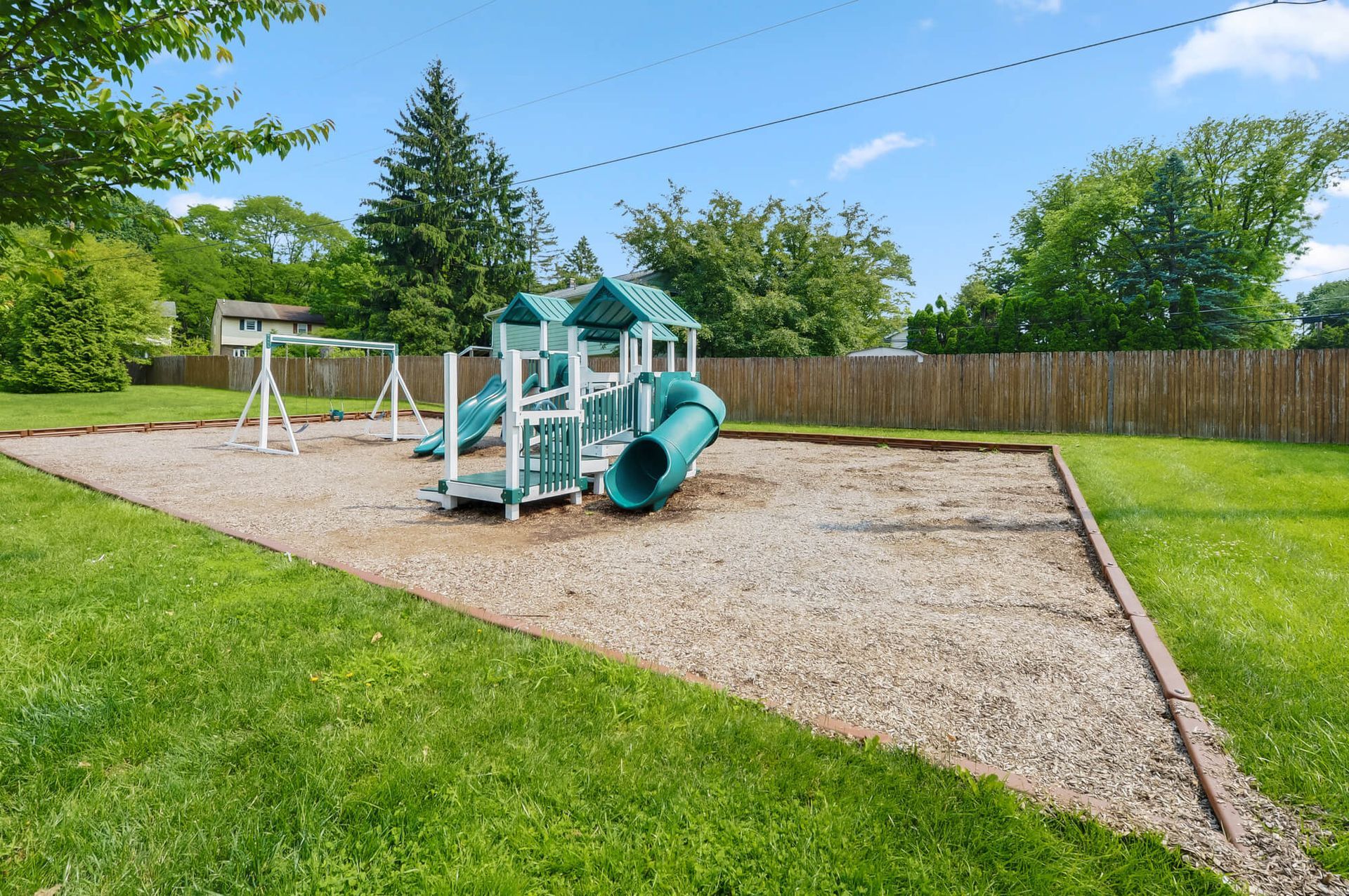 Playground with teal slides and swings on a gravel area, fenced by a wooden barrier.