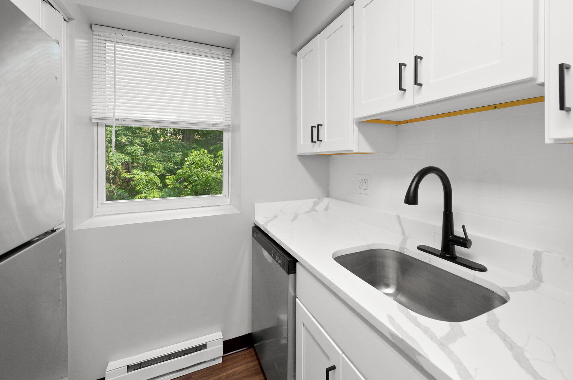 White kitchen with marble countertop, black faucet, stainless sink, and a window with greenery outside.