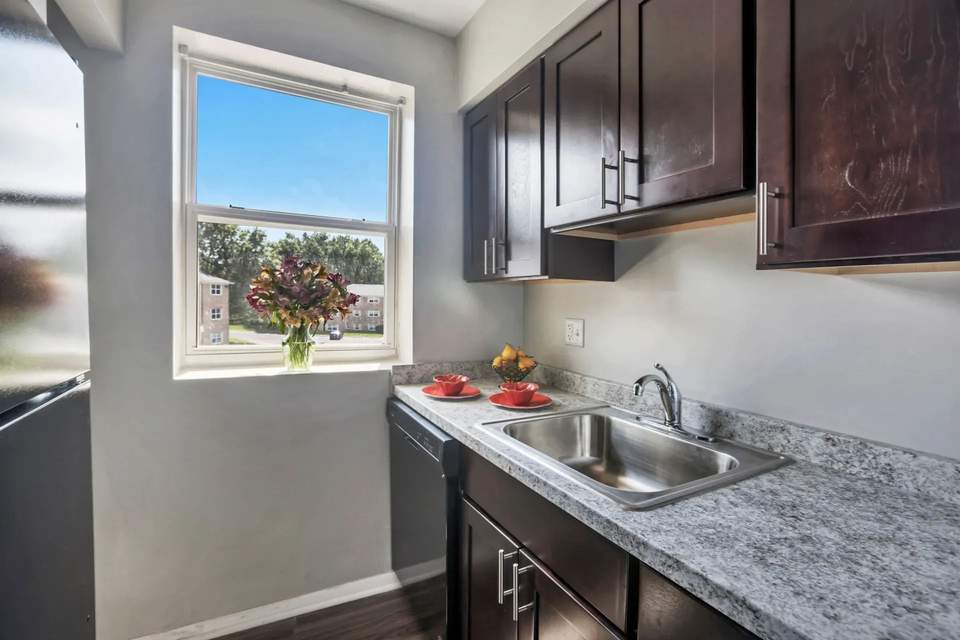 Kitchen in an apartment with dark wood cabinets, granite countertops, stainless sink, and a sunny window.