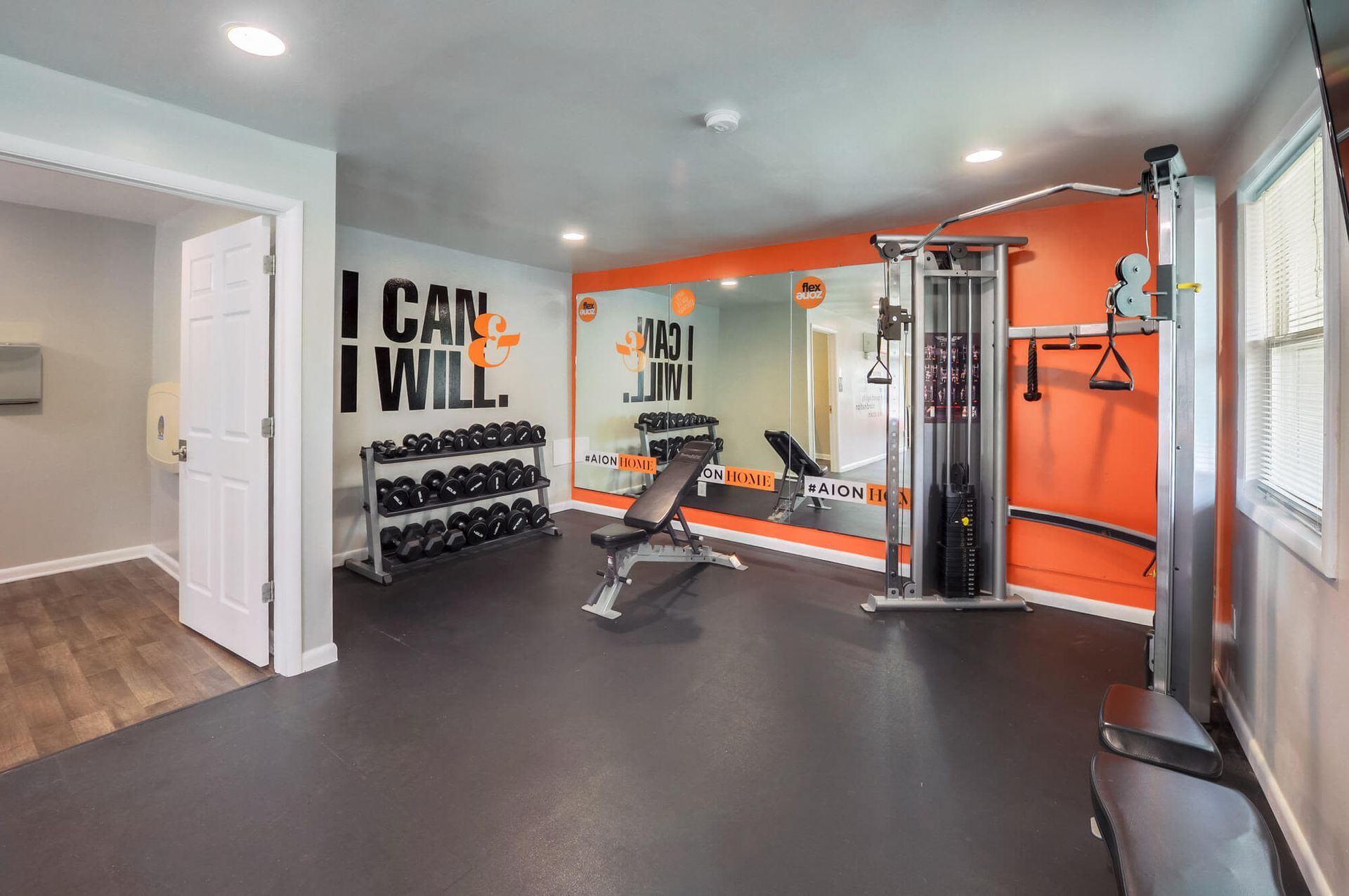 Interior of a resident gym with a weight rack, bench, and cable machine against an orange wall.