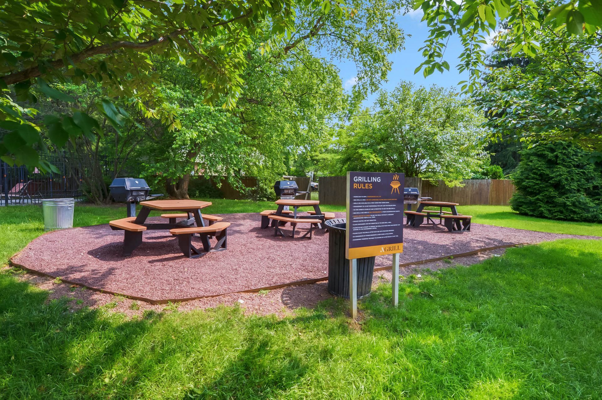 Outdoor community grilling area with wooden picnic tables and a sign in a tree-filled yard.