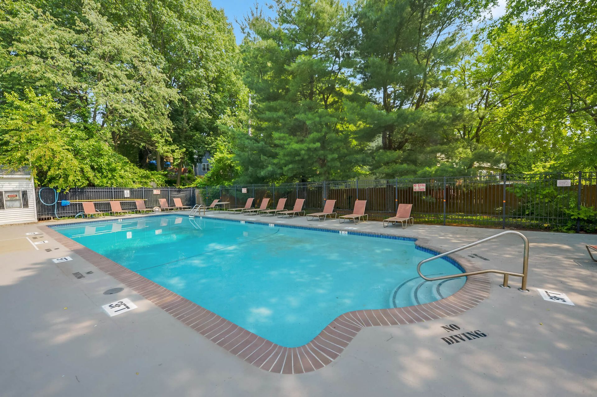 Outdoor apartment pool with blue water, lounge chairs, and green trees nearby.