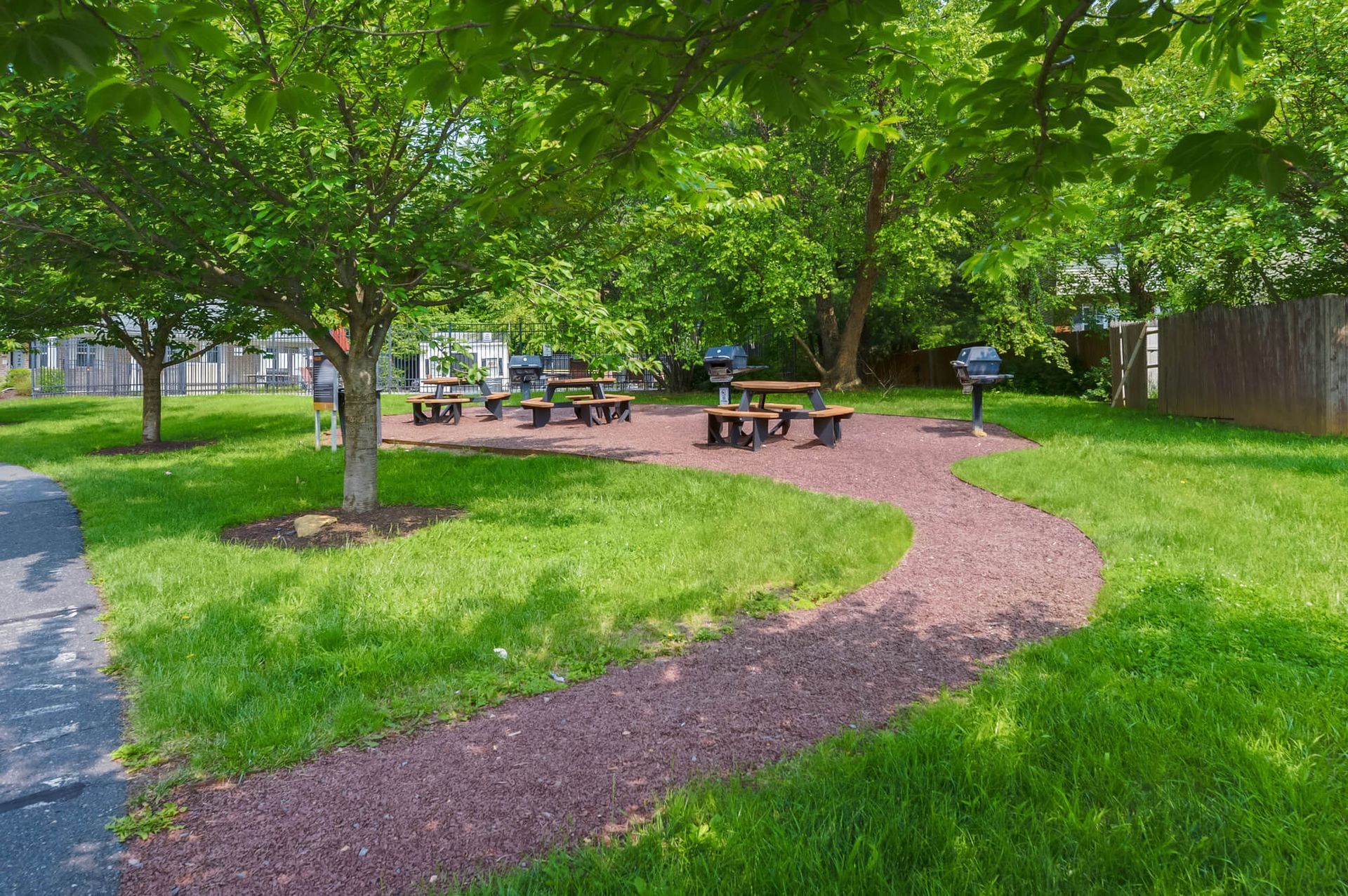 Outdoor community area with picnic tables, grills, trees, and a curved mulch path.