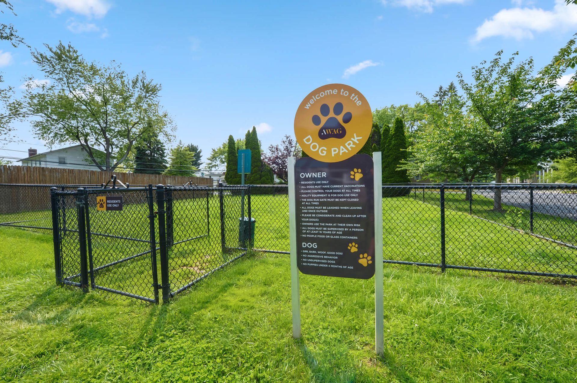 Dog park area with a round yellow welcome sign and a rules board inside a chain-link fence.
