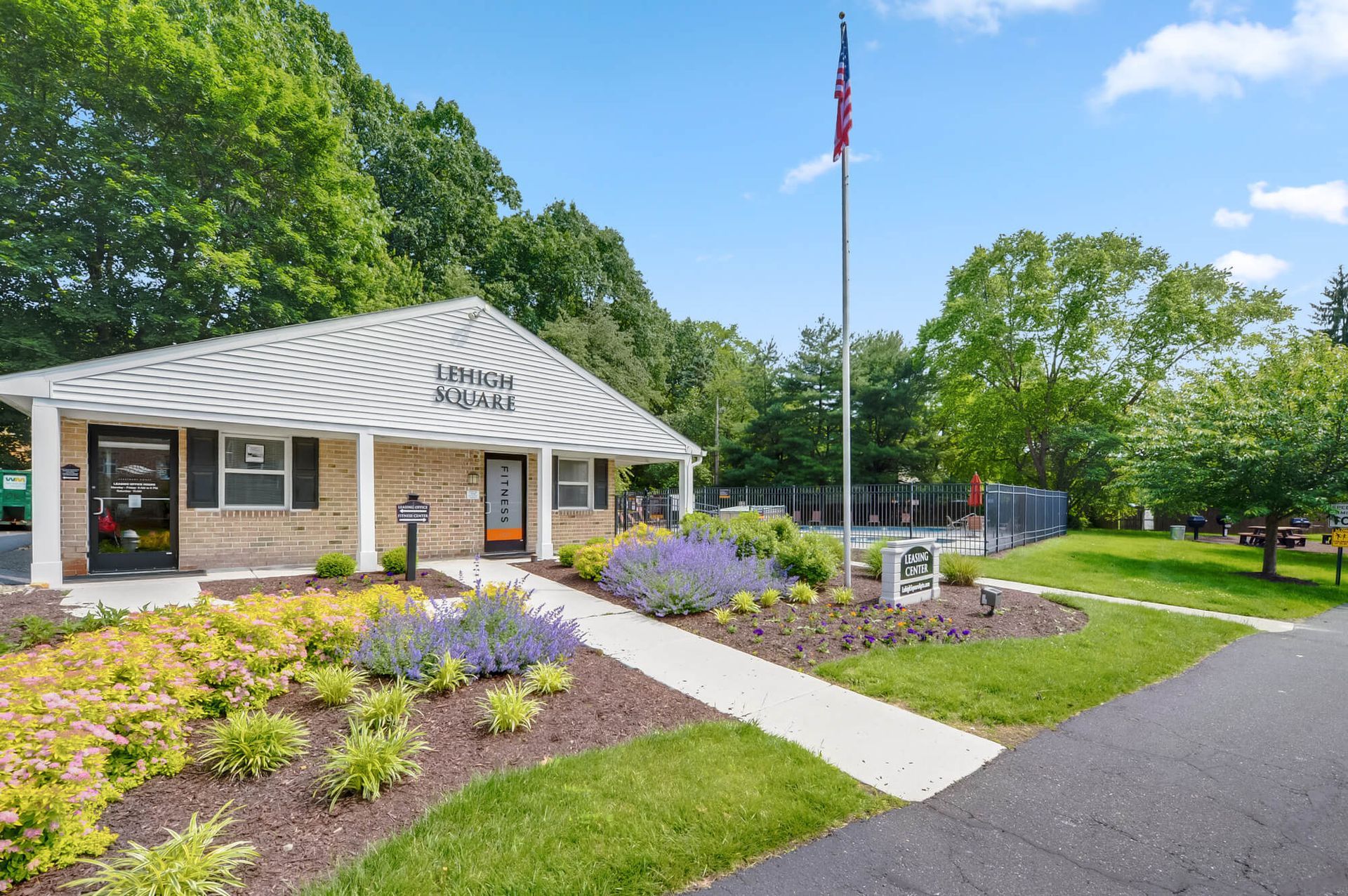 Exterior view of Lehigh Square leasing center with landscaped entrance, flagpole, and a fenced amenity area.