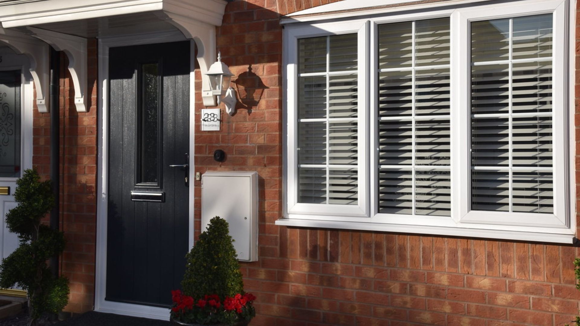 Front porch of a brick house with a black door, white shutters, and potted shrubs.