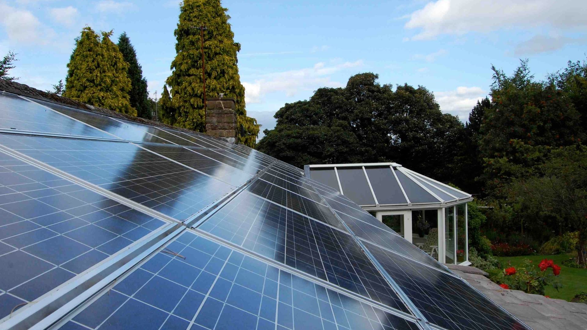 Solar panels on a rooftop in a sunny garden, with trees and a small building in the background.