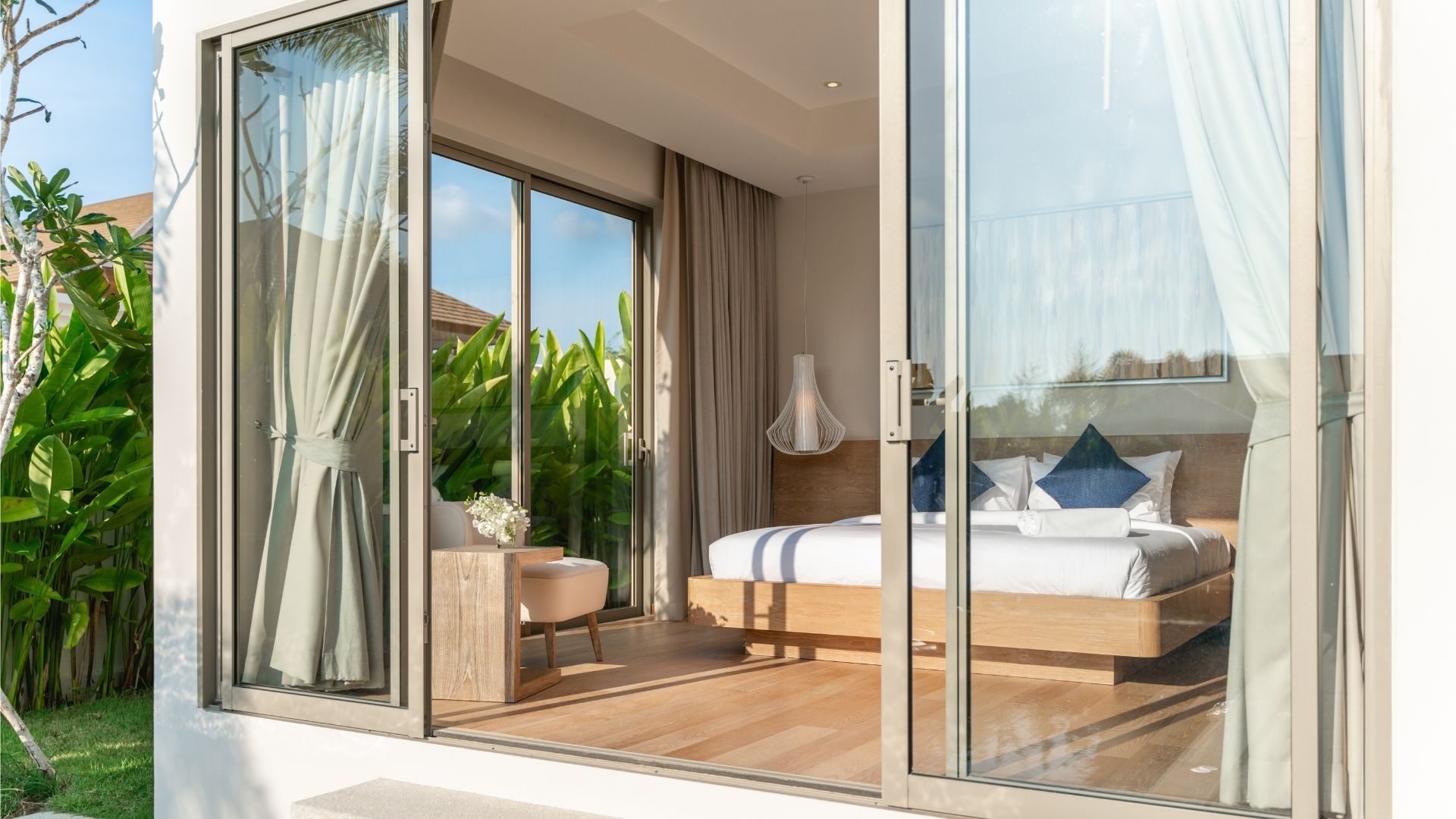 Open glass doors to a bright bedroom with a bed, lounge chair, and tropical outdoor view