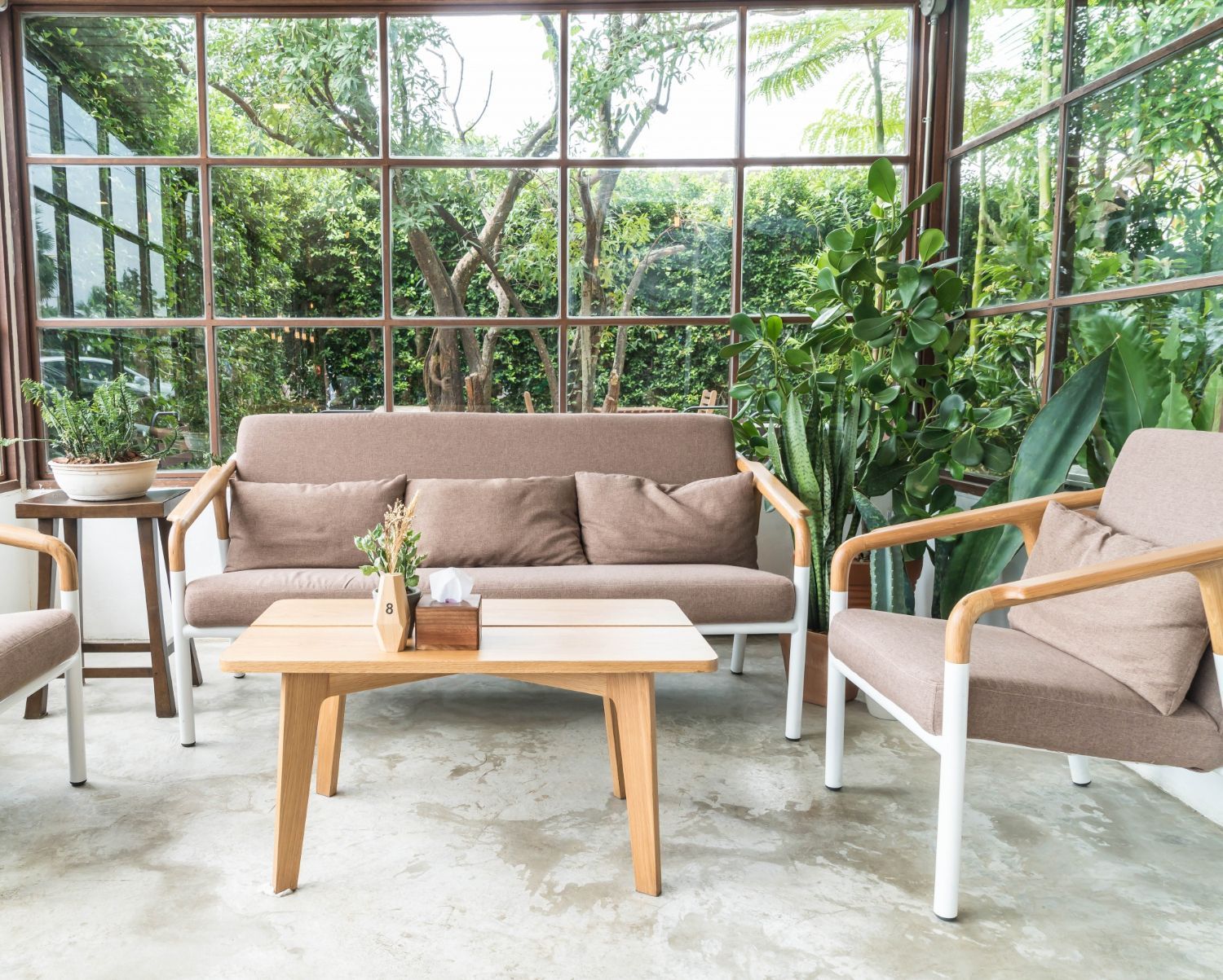Modern patio seating area with beige sofas, wooden tables, and lush green plants beside large windows