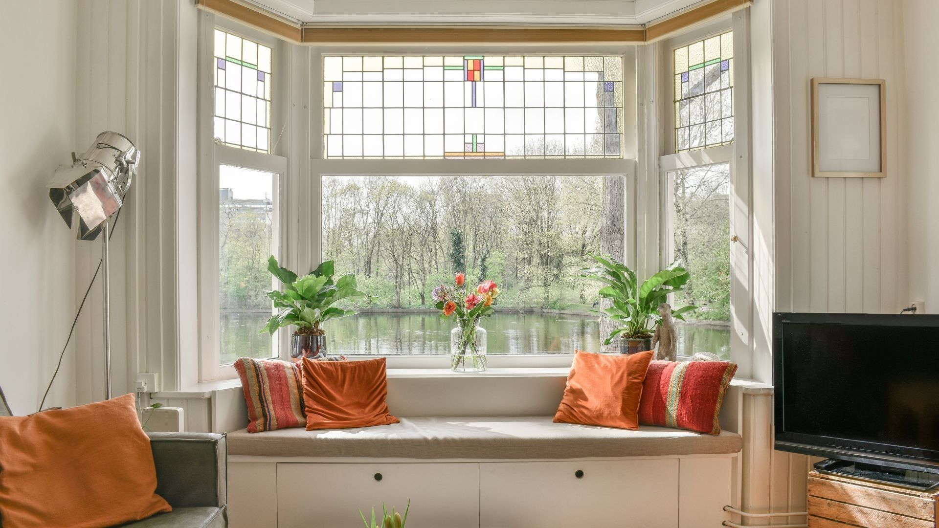 Bright bay window seat with orange cushions, potted plants, and a TV in a cozy living room