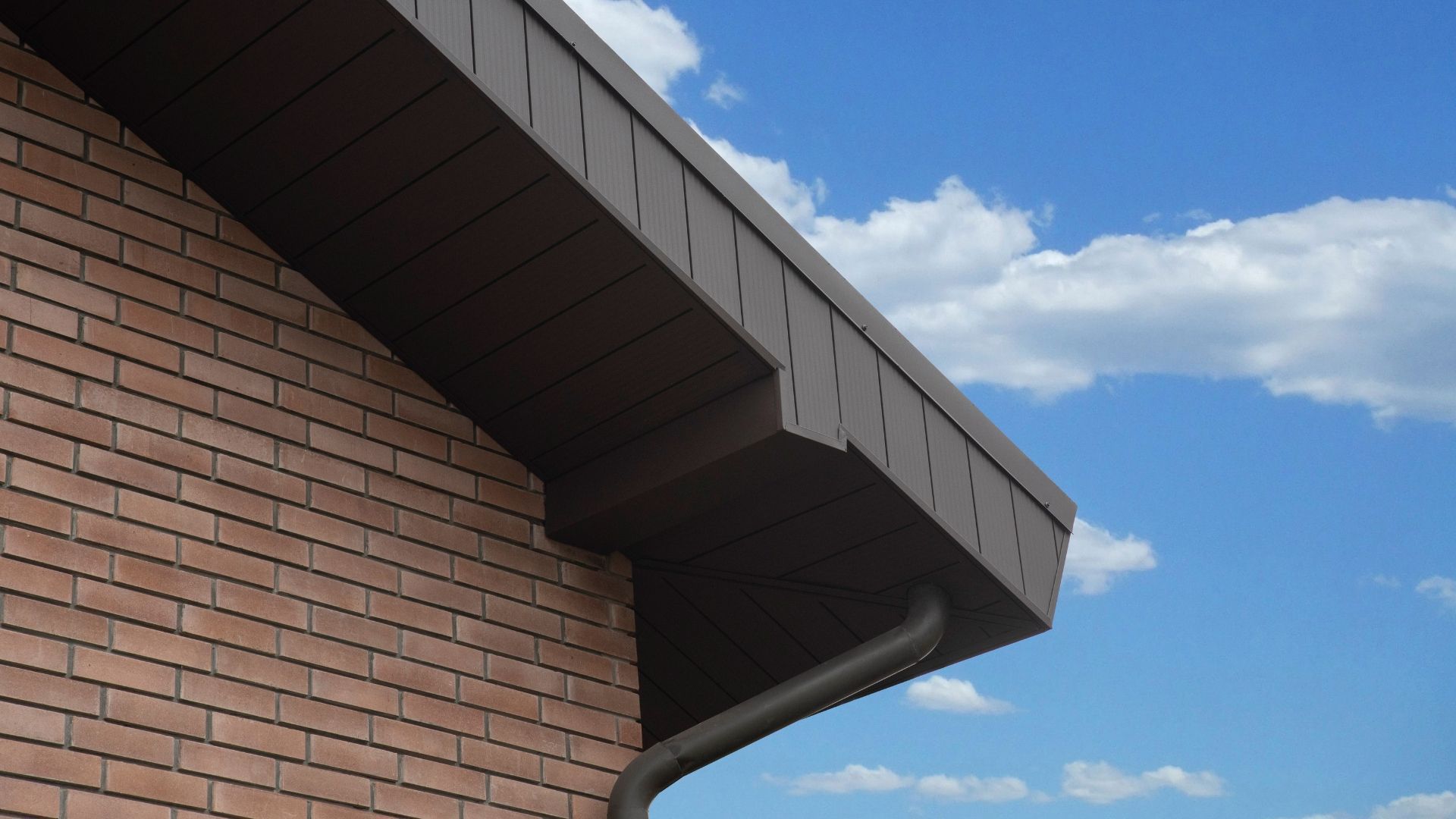 Brick building corner with dark roof overhang against a blue sky with clouds