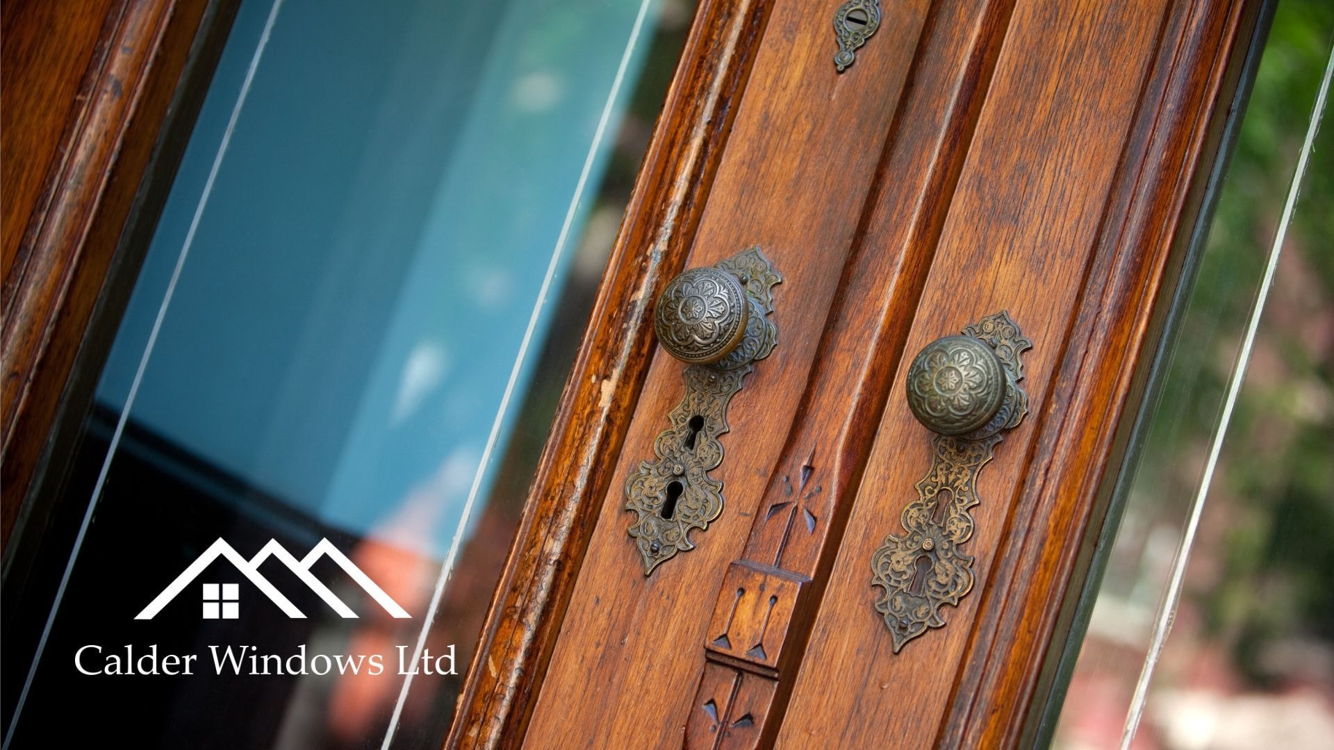 Close-up of a wooden door with ornate metal knobs and a window reflecting blue sky.