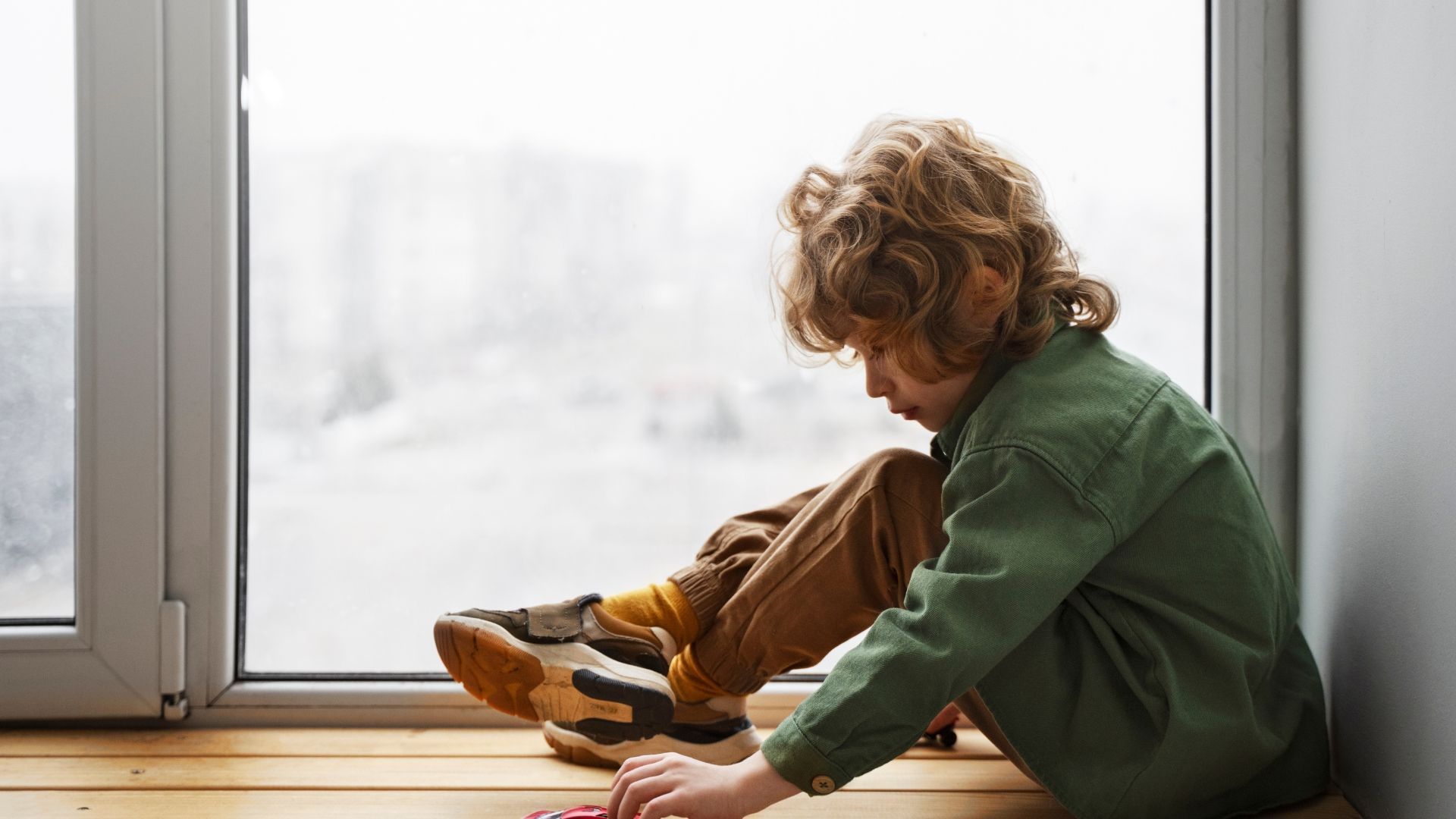 Child in a green sweater sits by a window, tying shoes on a wooden floor.