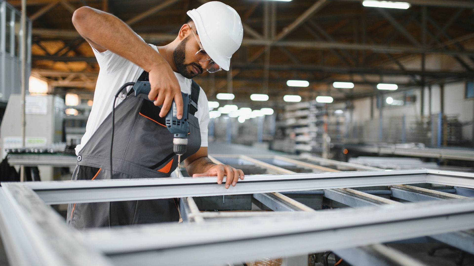 Worker in hard hat using a power drill on a metal frame in a factory workshop.