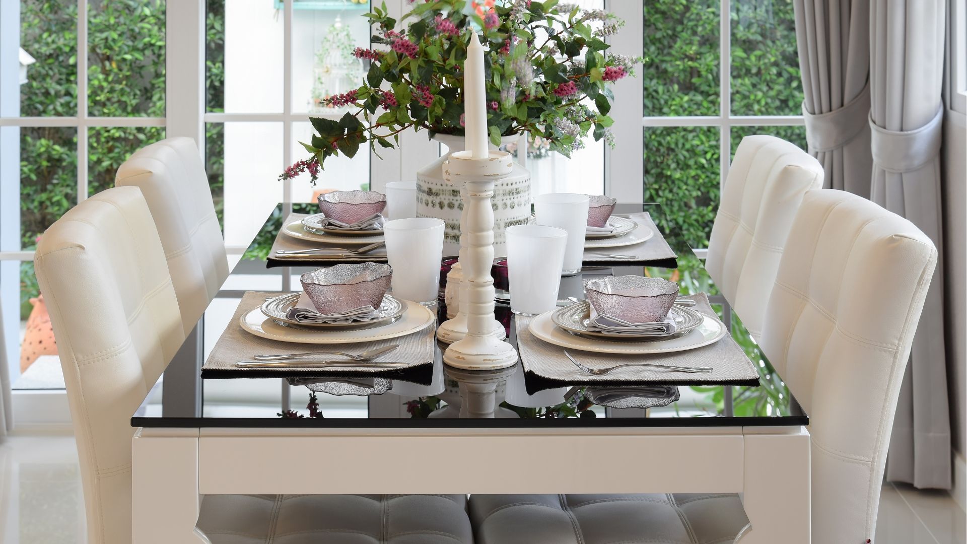 Elegant dining table set with cream chairs, glass top, white floral centerpiece, and place settings by a bright window